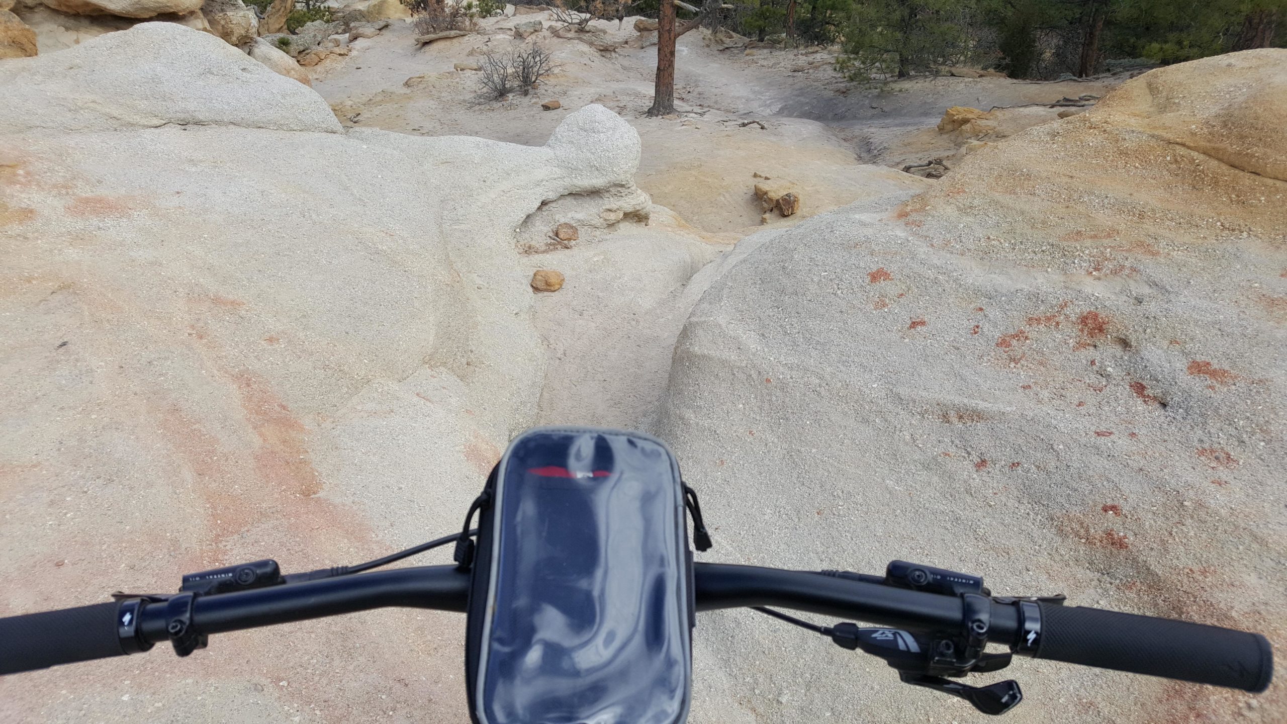 A view from the handlebars of a mountain bike overlooking a rocky, uneven terrain with light-colored stone and patches of vegetation in the background. Palmer Park mountain bike trail.