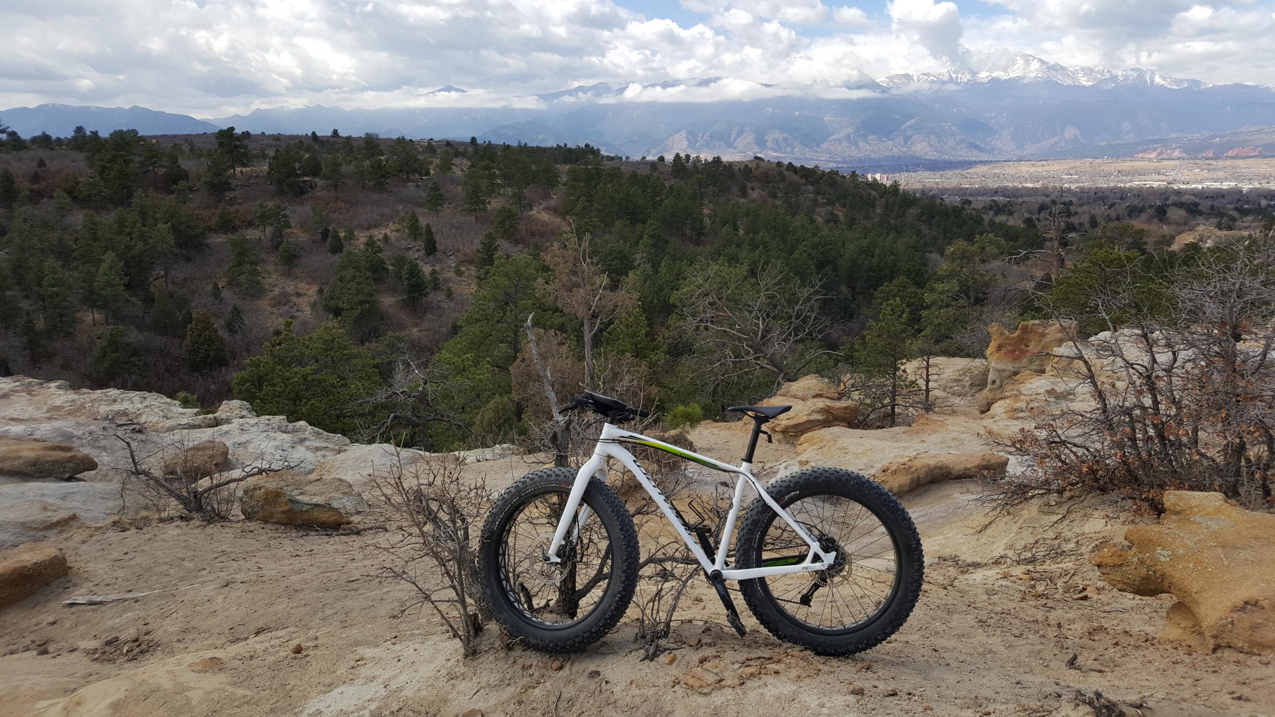 A white fat bike is leaning against a rocky outcrop, set against a backdrop of rolling hills and pine trees. In the distance, snow-capped mountains can be seen under a partially cloudy sky. The terrain is sandy with patches of dry vegetation, creating a rugged outdoor scene. Palmer Park mountain bike trail.