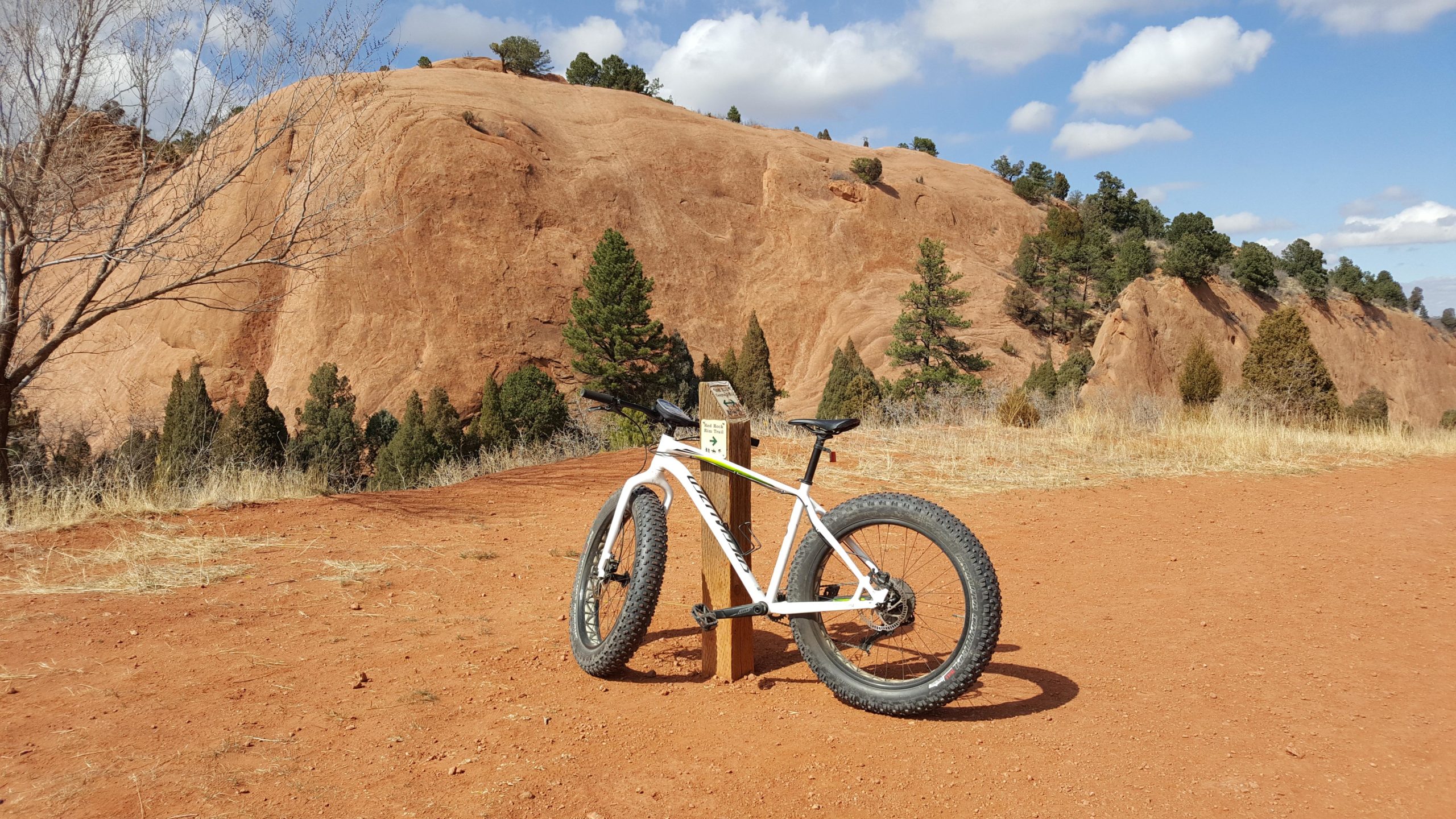 A fat tire bicycle leaned against a wooden signpost on a dirt trail, with red rock formations and green trees in the background under a partly cloudy sky. Red Rock Canyon mountain bike trail.