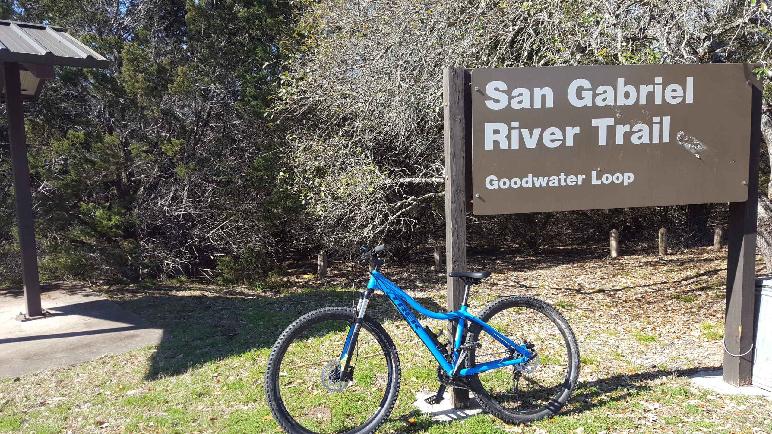 Alt tag: A blue mountain bike is parked next to a sign for the San Gabriel River Trail, Goodwater Loop, surrounded by green trees and a grassy area. Goodwater Trail mountain bike trail.