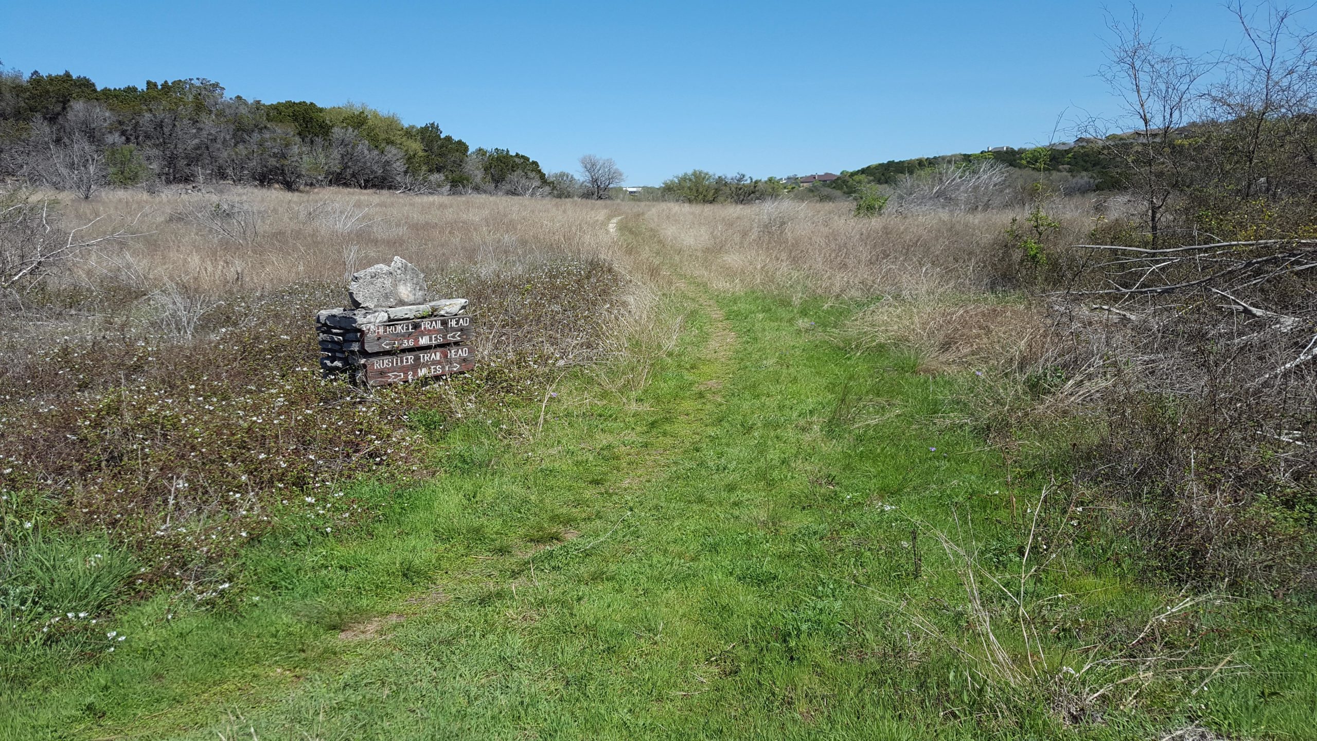 A scenic outdoor pathway leading through a grassy field, with a wooden sign indicating trail head distances for hiking, against a clear blue sky. The trail is flanked by sparse vegetation and trees in the background. Goodwater Trail mountain bike trail.