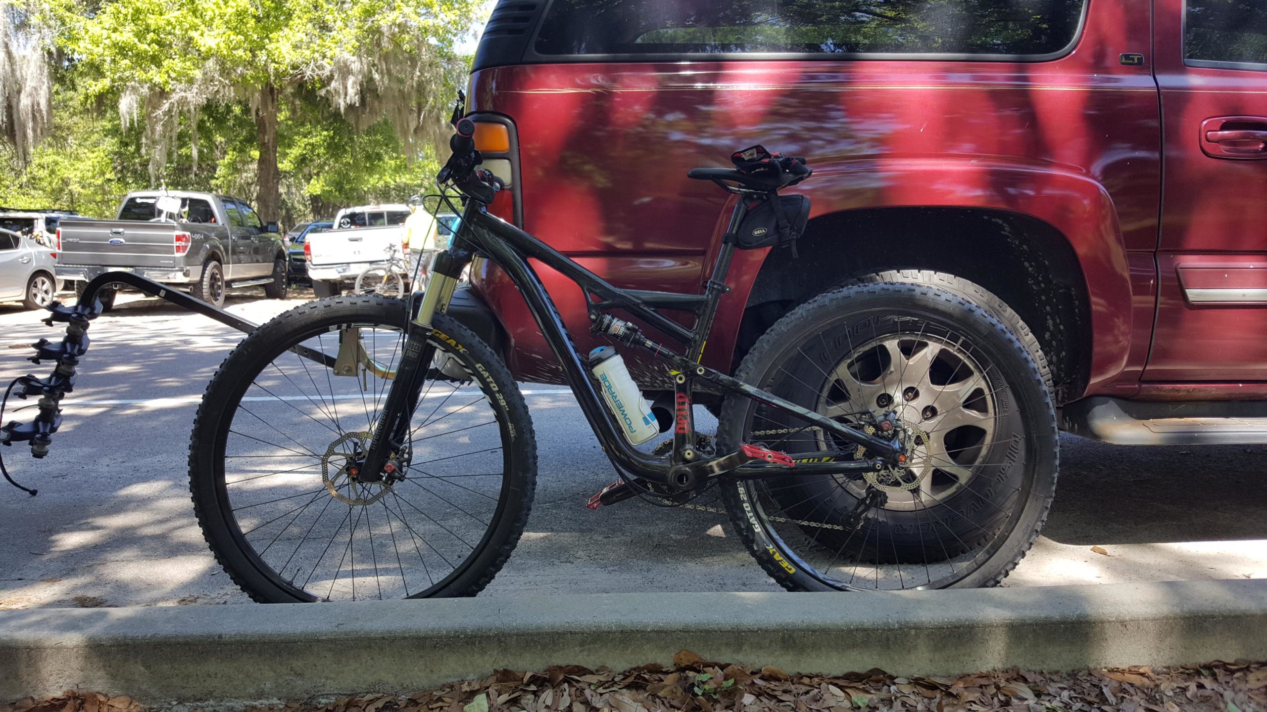 A black mountain bike is parked on a concrete curb next to a red pickup truck in a shaded area. The bike has thick tires and a water bottle attached to its frame. Various parked vehicles can be seen in the background, along with green trees above. Alafia River State Park mountain bike trail.