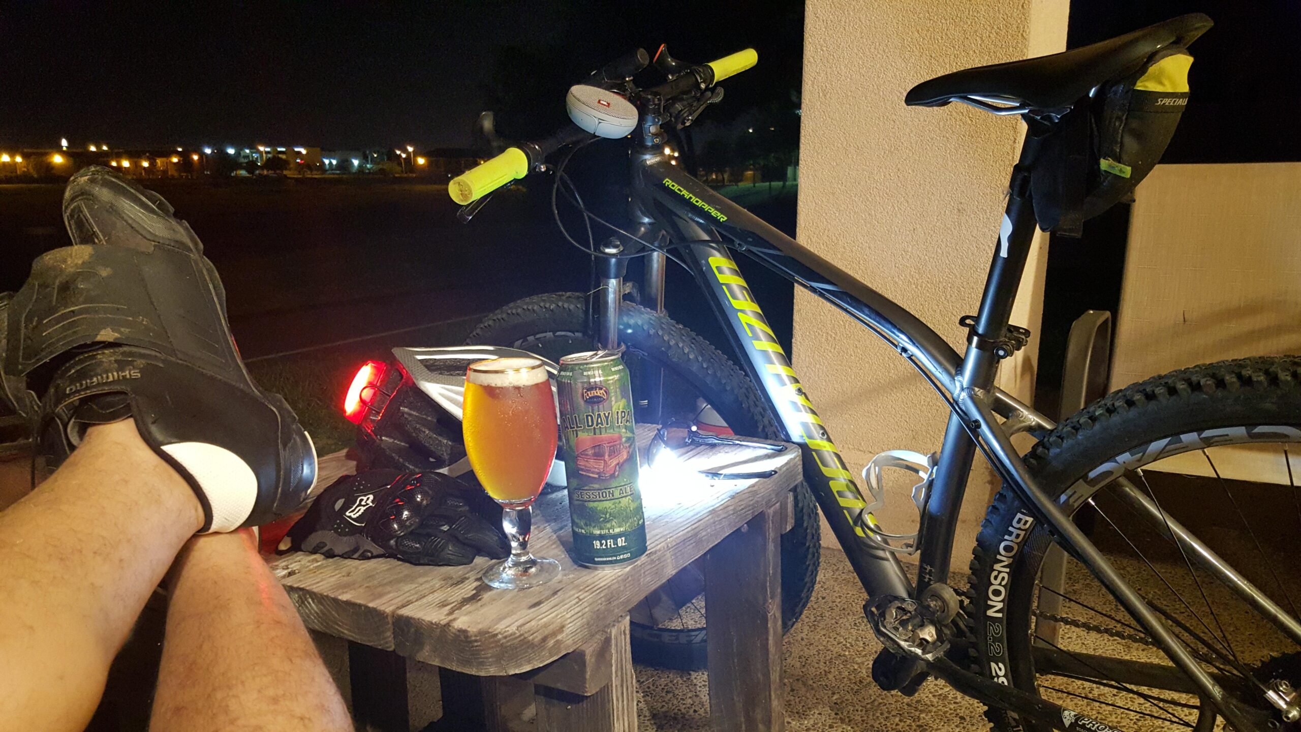 Specialized Rockhopper Comp 29: A pair of feet resting on a wooden table, wearing cycling shoes, with a mountain bike nearby. A glass of beer and a can of "All Day IPA" are on the table, accompanied by a bike helmet and gloves. The scene is illuminated by soft lighting at night, with blurred city lights in the background.