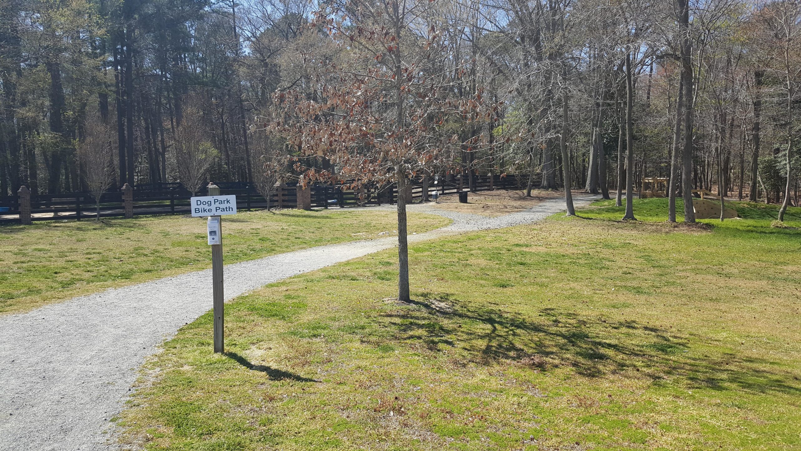 A winding gravel pathway leading through a grassy area, with a sign indicating "Dog Park Bike Path" nearby. Lush green grass and scattered trees are visible, with a wooded area in the background. The scene is set in a bright, sunny environment, suggesting a peaceful outdoor space for dog walking and biking. Windsor Castle Park mountain bike trail.