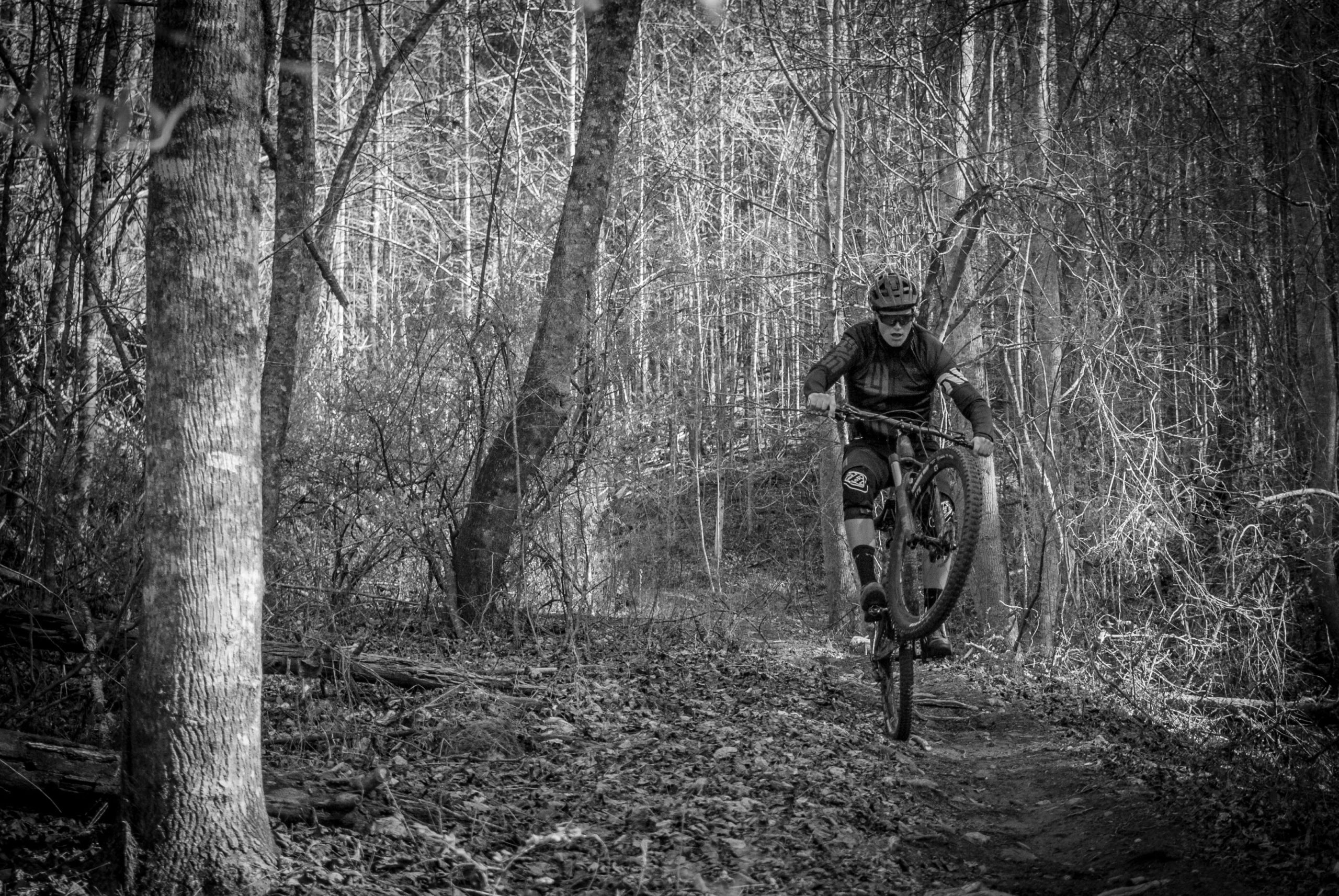 A mountain biker performing a wheelie on a dirt trail surrounded by trees in a forest, captured in black and white. Pinhoti Trail: P2 mountain bike trail.