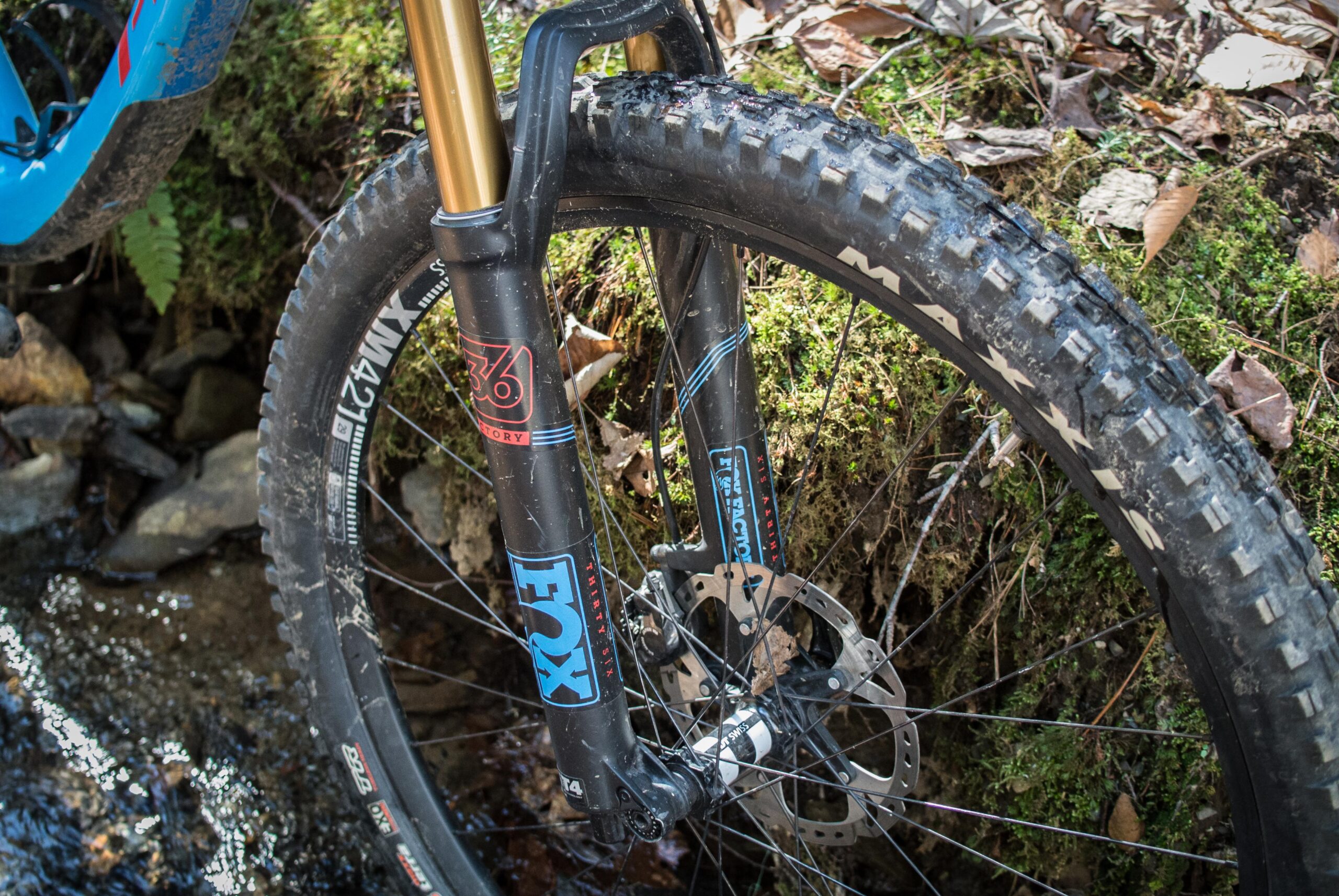 Pivot SwitchBlade: A close-up view of a mountain bike's front wheel and fork, featuring a black suspension fork with gold stanchions, resting on a mossy bank next to a shallow stream. The wheel has a rugged, heavily treaded tire with "Maxxis" branding, and a disc brake is visible alongside the spokes. Surrounding foliage includes dry leaves and green moss, suggesting an outdoor trail environment.