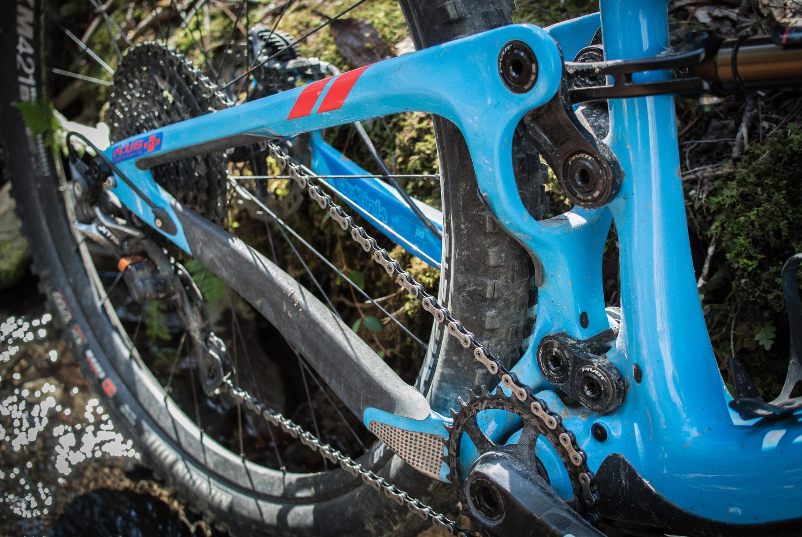 Pivot SwitchBlade: Close-up view of a blue mountain bike frame showcasing the rear triangle, chain, and drivetrain components. The image highlights the bike's suspension linkage and tire, set against a natural background with some greenery.