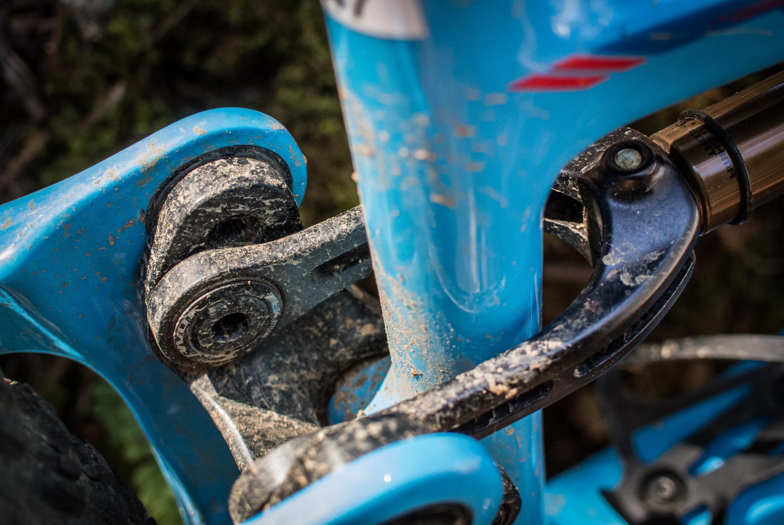 Pivot SwitchBlade: Close-up view of a blue mountain bike's suspension linkage, showing dirt and wear on the components, with a focus on the pivot points and lower frame structure.