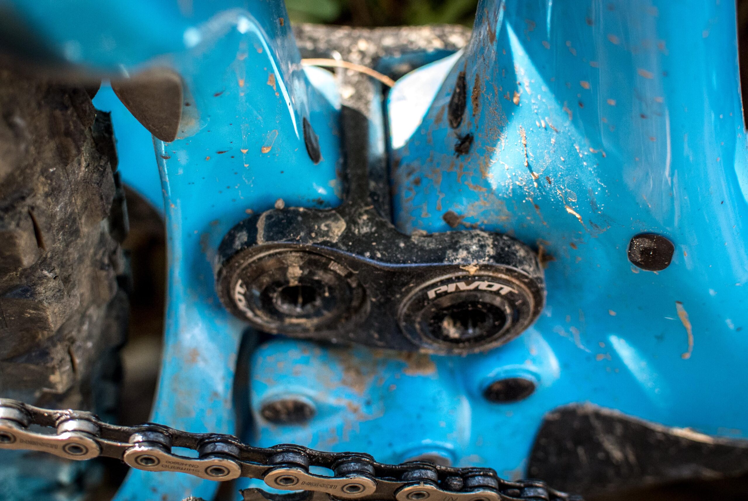 Pivot SwitchBlade: Close-up view of a blue bicycle frame featuring the bottom bracket area, showing a black pivot system and a portion of the chain. The frame is slightly muddy, suggesting recent use on rough terrain.