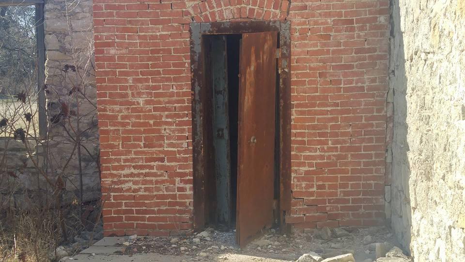 An old, rusty door partially ajar in a brick wall, leading to a dark interior. The brickwork is weathered and shows signs of age, with some exposed bricks and a cracked foundation. Surrounding vegetation and debris are evident on the ground. Flint Hills Nature Trail (Rail Trail) mountain bike trail.