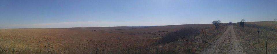 A panoramic view of expansive golden grasslands under a clear blue sky, featuring a dirt road winding through the landscape. Sparse trees are visible on the horizon, enhancing the serene rural atmosphere. Flint Hills Nature Trail (Rail Trail) mountain bike trail.