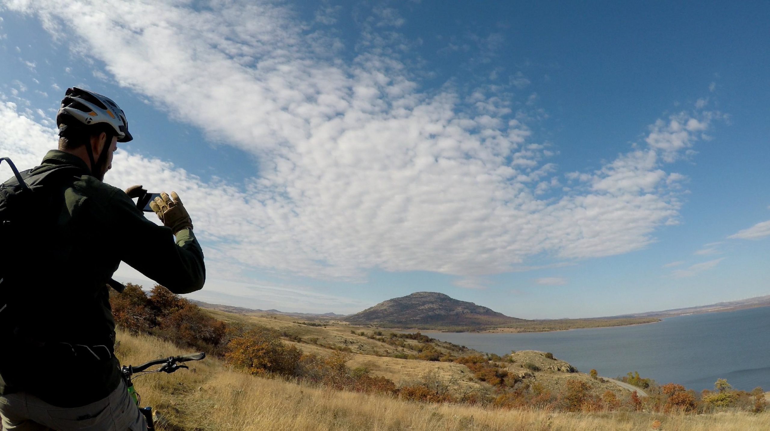 A person wearing a helmet and gloves is standing on a grassy hillside, taking a photo with a smartphone. In the background, a serene lake is visible, flanked by rolling hills and a prominent mountain. The sky is bright blue with scattered clouds, and colorful foliage can be seen in the landscape. Lake Lawtonka Trails mountain bike trail.