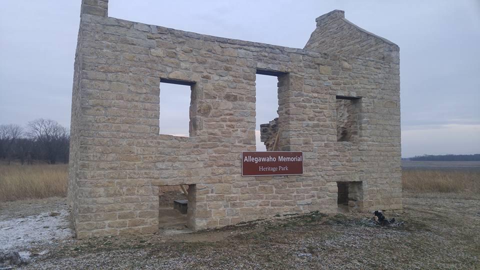 A weathered stone building with partially collapsed walls, featuring a sign that reads "Allegawaho Memorial Heritage Park." The structure is set against a cloudy sky and surrounded by tall grass, indicating a rural landscape. Flint Hills Nature Trail (Rail Trail) mountain bike trail.