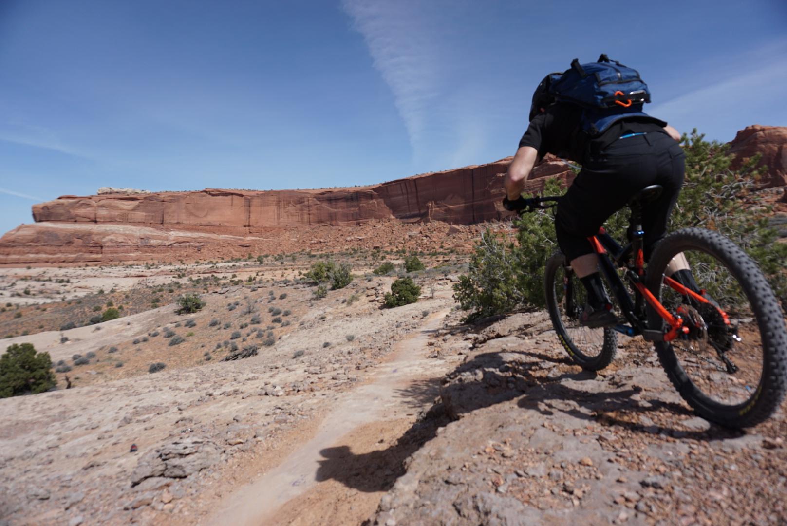A mountain biker navigating a rocky trail with red rock formations in the background under a clear blue sky. Navajo Rocks mountain bike trail.