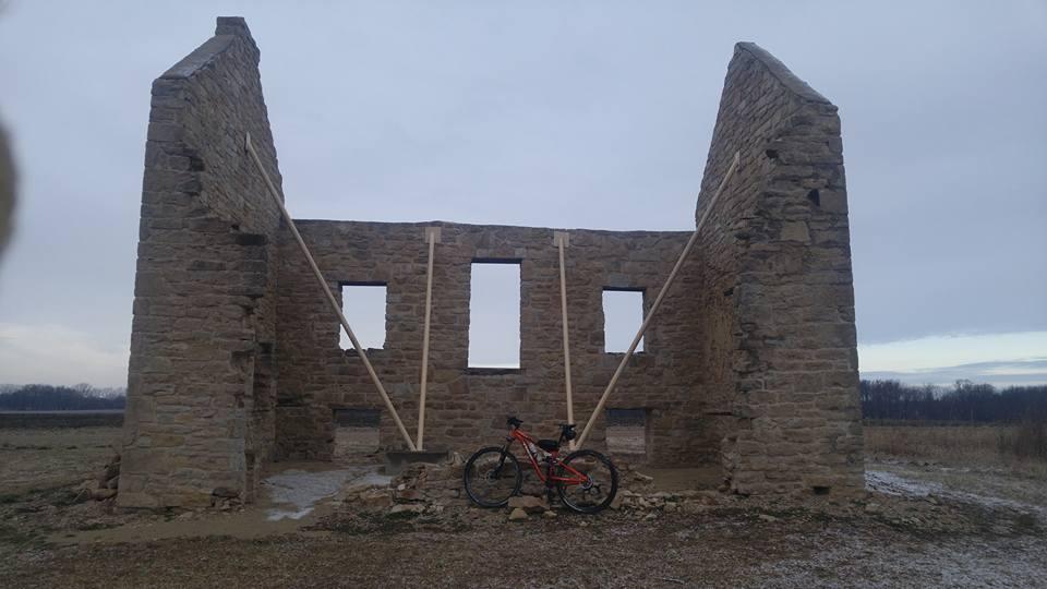 Alt text: A partially standing stone structure with visible support beams, set in an open field with a cloudy sky. A mountain bike is leaning against the wall in the foreground. Flint Hills Nature Trail (Rail Trail) mountain bike trail.