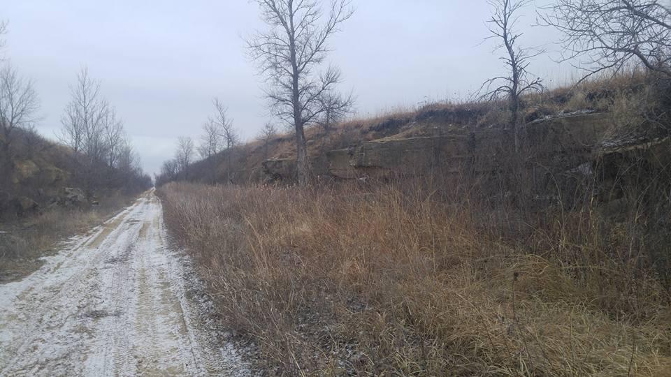 A dirt path runs through a landscape with sparse trees on either side, surrounded by tall grasses and rocky outcroppings. The scene is set under a cloudy sky, indicating a cool, overcast day. Snow is visible along the path, suggesting recent winter weather. Flint Hills Nature Trail (Rail Trail) mountain bike trail.