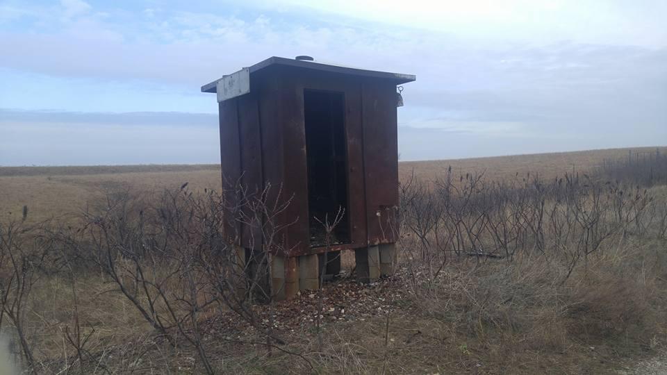 A small, rustic wooden shed with a brown exterior stands on stilts in a barren landscape, surrounded by sparse vegetation. The sky is overcast, creating a moody atmosphere, while the open door of the shed suggests it is empty or abandoned. Flint Hills Nature Trail (Rail Trail) mountain bike trail.