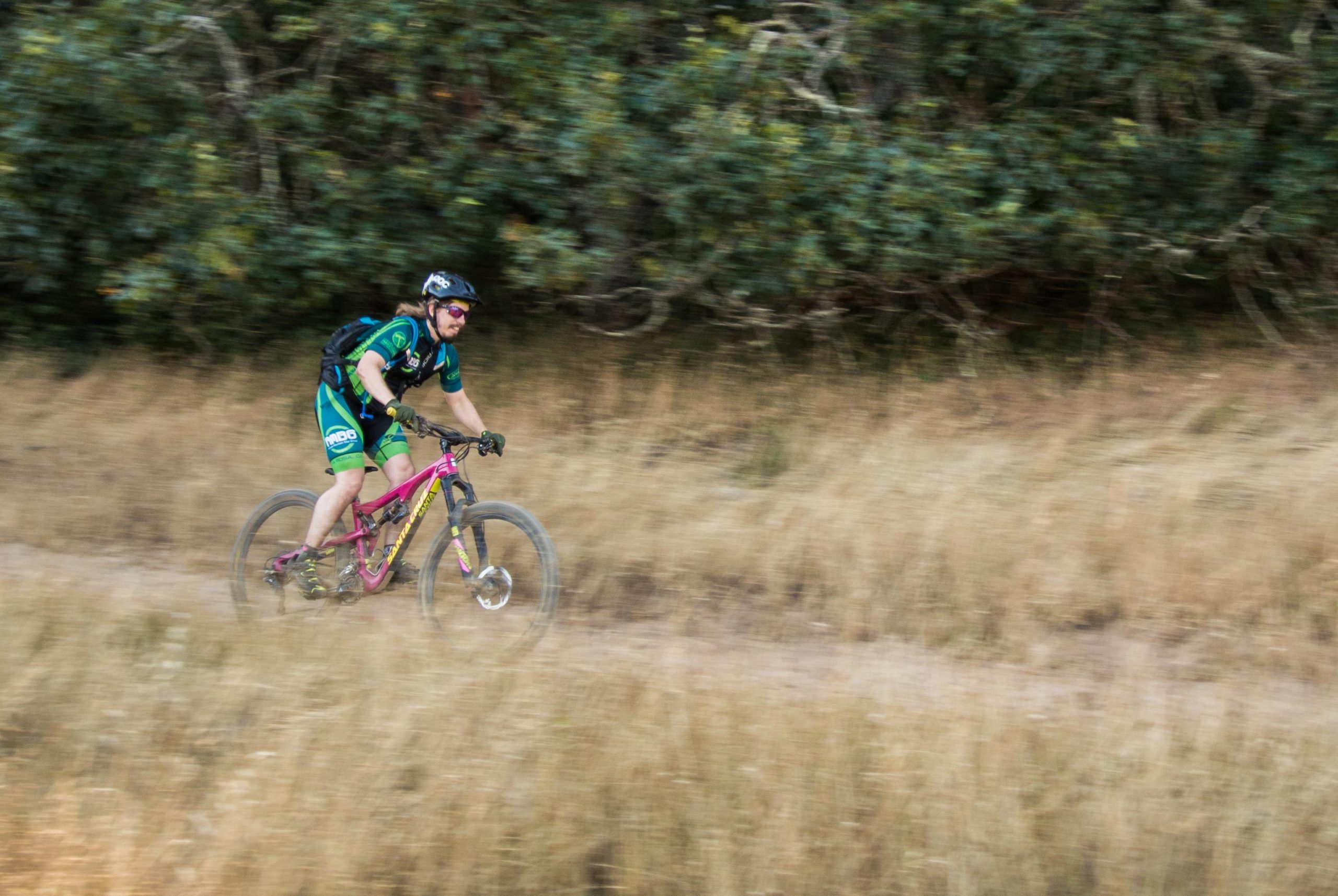 A mountain biker in a green and blue jersey rides swiftly along a dirt trail, surrounded by tall grass and shrubs. The motion suggests speed, with a blurred background indicating movement. The biker wears a helmet and sunglasses for protection. Annadel State Park mountain bike trail.