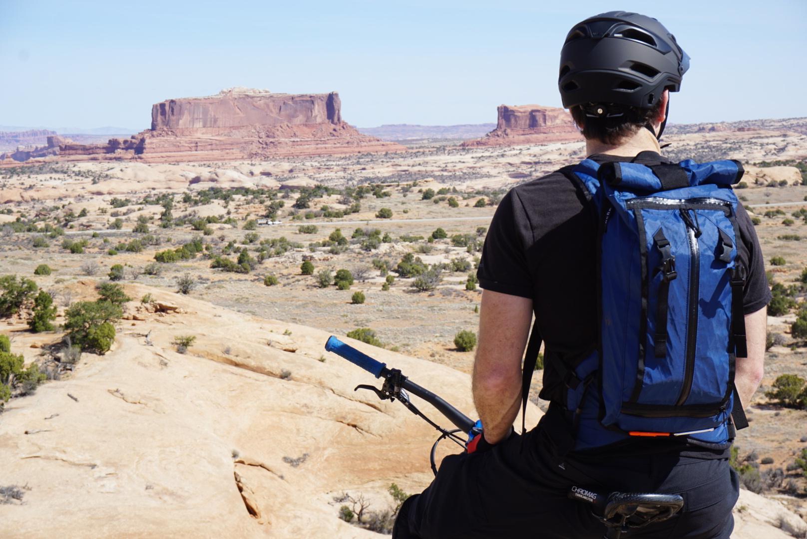 A person wearing a black helmet and a blue backpack sits on a rocky ledge, gazing at a vast desert landscape with mesas and sparse vegetation under a clear blue sky. They hold a mountain bike handlebar, suggesting they are a cyclist enjoying the scenic view. Navajo Rocks mountain bike trail.