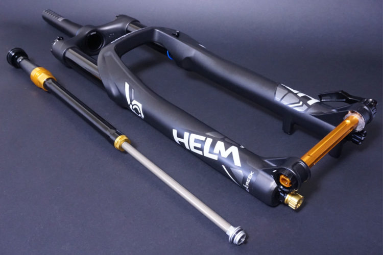 A close-up of a mountain bike fork, featuring a black frame with white branding and orange accents. The fork is displayed alongside a removable suspension cartridge, showcasing its design and components against a dark background.