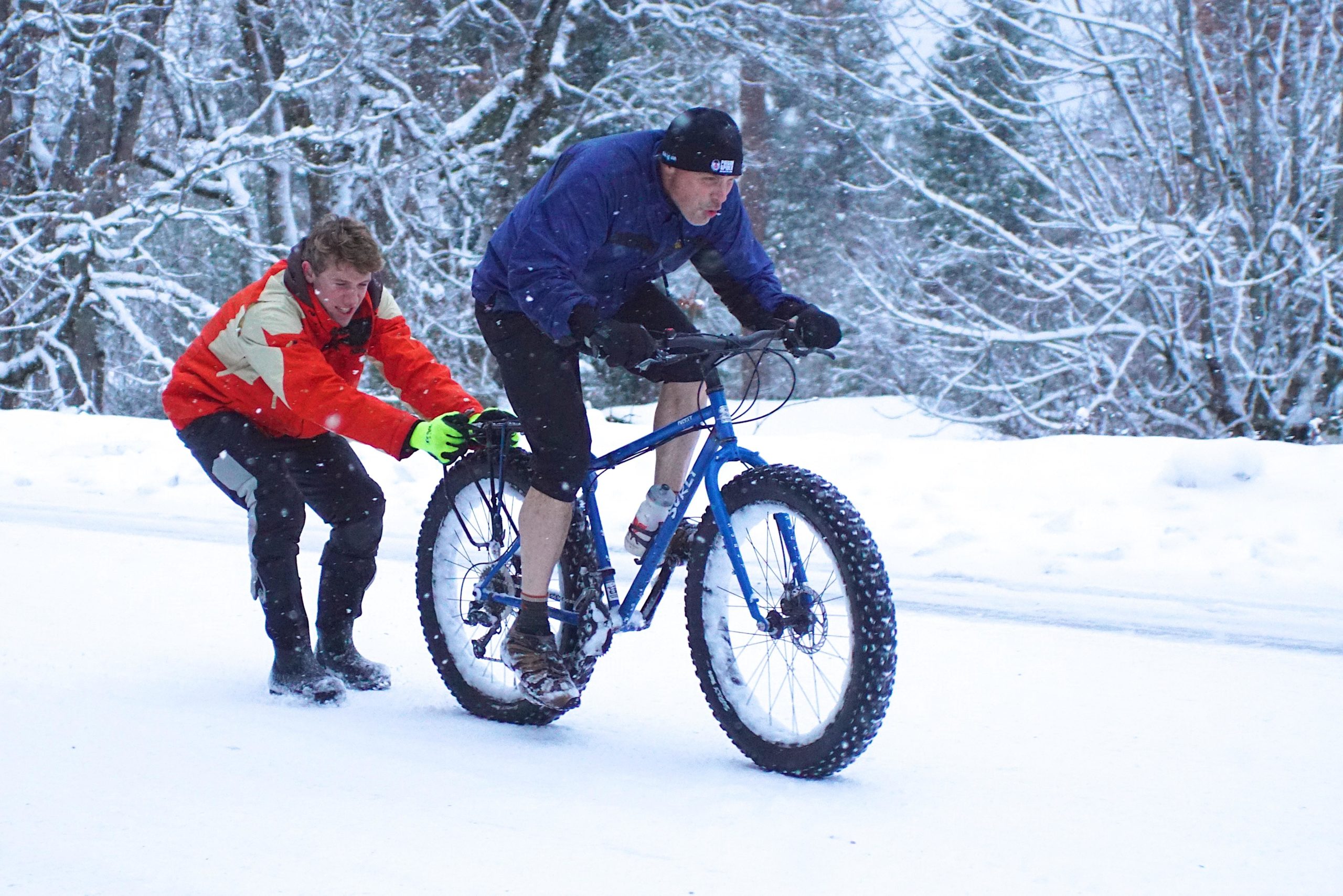 A person in a red and beige jacket pushes a cyclist riding a blue fat bike on a snowy road. Snowflakes are falling, and the landscape is covered in a thick layer of snow, with trees in the background. The cyclist is focused, wearing a hat and shorts, while the helper looks determined as he assists in the effort. Syncline mountain bike trail.