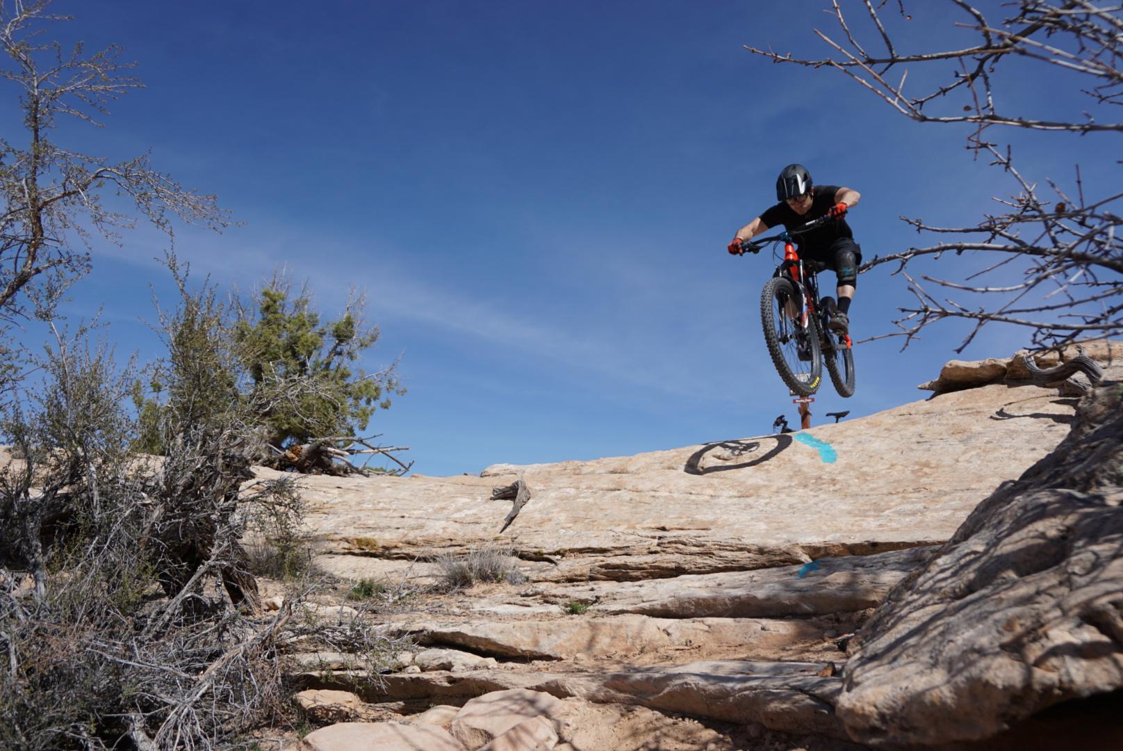 A mountain biker performing a jump over a rocky terrain under a clear blue sky, with sparse vegetation in the foreground. Navajo Rocks mountain bike trail.