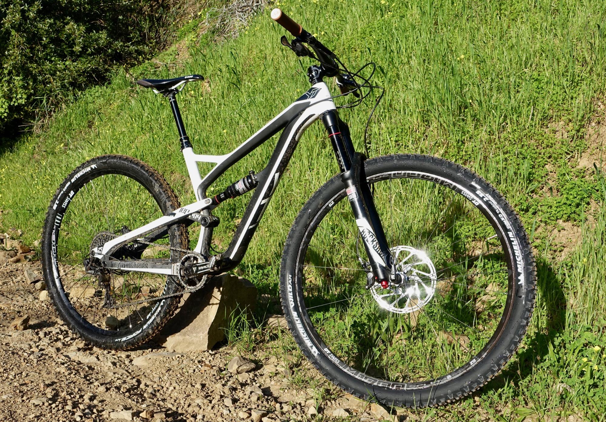 Yt industries Jeffsy: A mountain bike leaning against a rock on a gravel trail, surrounded by lush green grass. The bike features a black and white frame, large tires with a rugged tread, and a textured handlebar grip. Sunlight highlights the details of the bike against the natural backdrop.