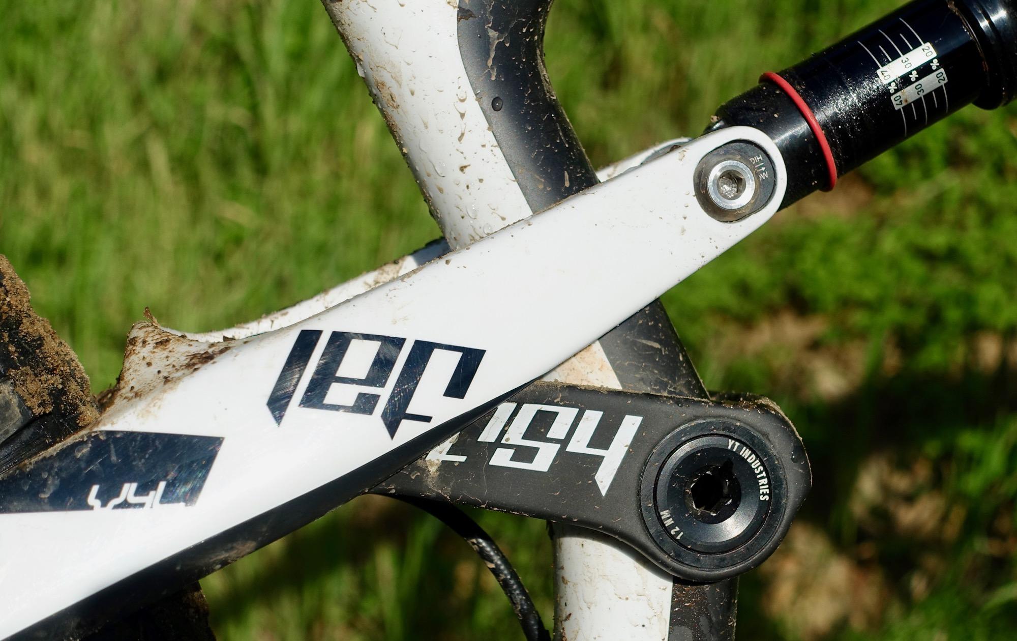 Yt industries Jeffsy: Close-up of a mountain bike's rear suspension arm, featuring a white frame with black decals. The arm is slightly muddy, highlighting its use in outdoor terrain. In the background, green grass is visible, indicating a natural environment.
