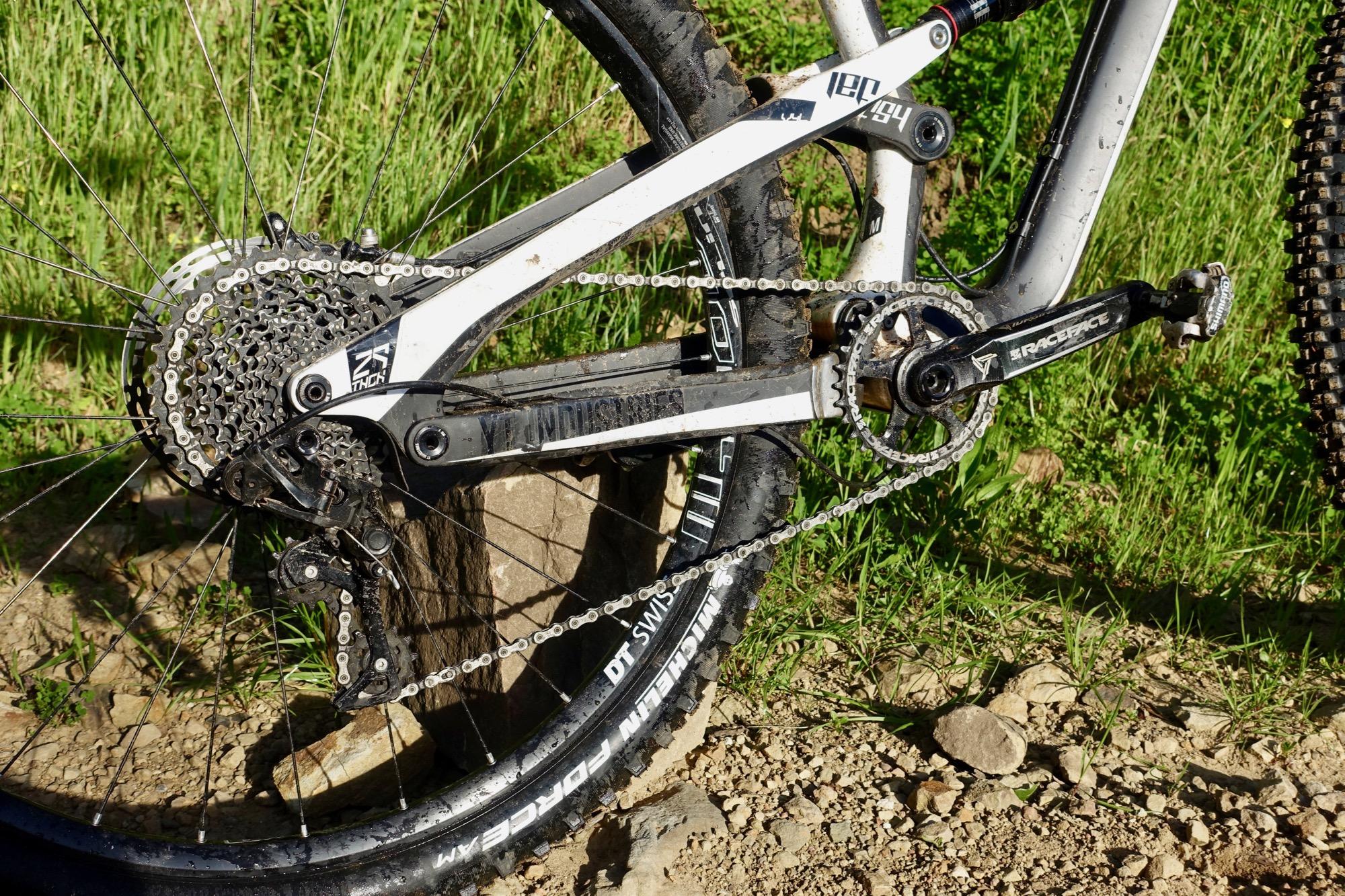 Yt industries Jeffsy: Close-up of a mountain bike's rear drivetrain, showcasing the gears, chain, and crankset. The bike has a white and black frame, with a visible derailleur and a muddy tire. The background features grass and pebbles, indicating an outdoor setting.
