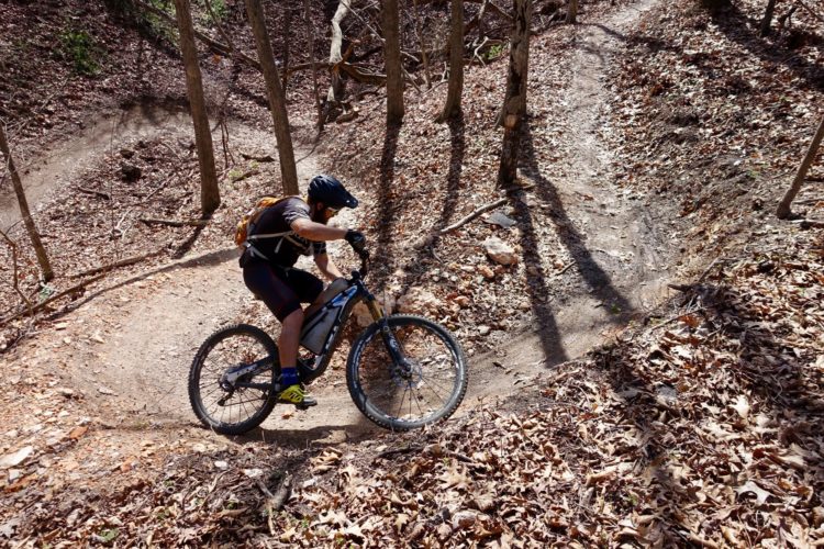 A person riding an electric mountain bike on a dirt trail in a forested area, surrounded by leaf-covered ground and trees. The rider is positioned with one foot on the ground and the other on the pedal, navigating a curved section of the trail.