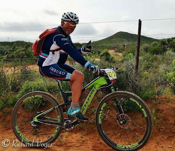 Cannondale Scalpel 29er 3: A mountain biker in a blue and white jersey poses with a thumbs-up while sitting on a green bicycle. He is wearing a helmet and has a backpack. The background features a dirt path and rolling green hills under a cloudy sky.