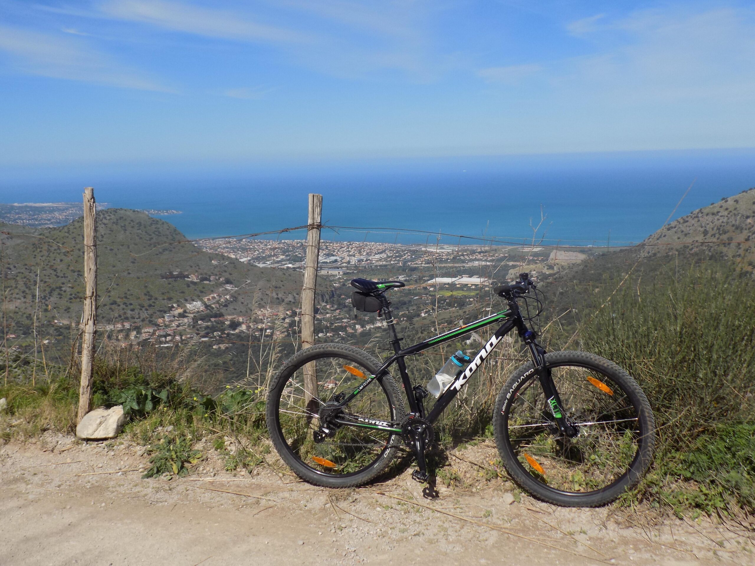 Kona Lava Dome: A mountain bike parked beside a dirt path with a scenic view of a coastal town and the sea in the background, under a clear blue sky.