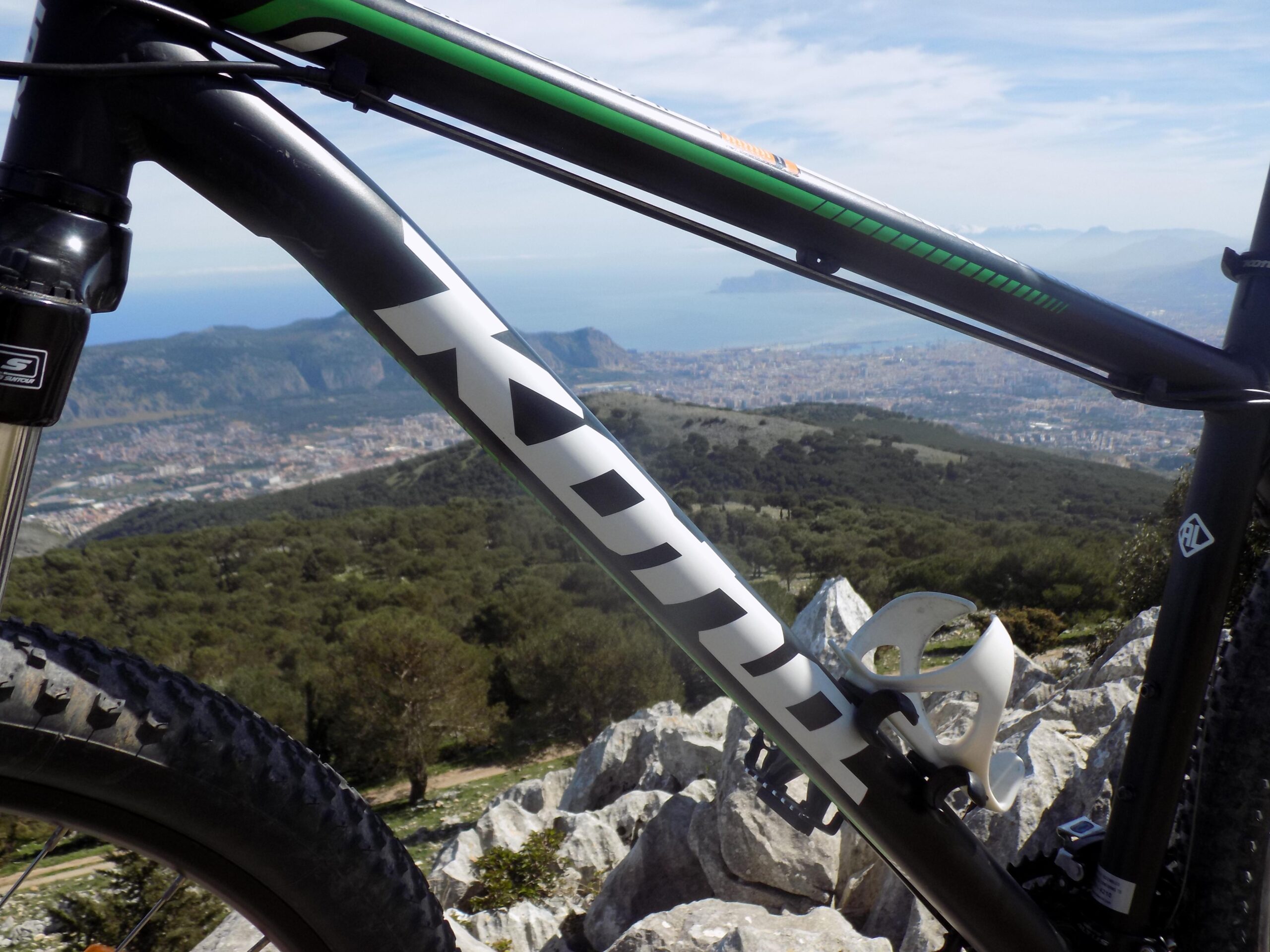 Kona Lava Dome: Close-up of a mountain bike frame with the brand name "Kona" visible, positioned against a scenic backdrop of hills and a coastal city. The image captures the bike's tire, part of the frame, and a water bottle holder, with rocky terrain and lush greenery in the background.