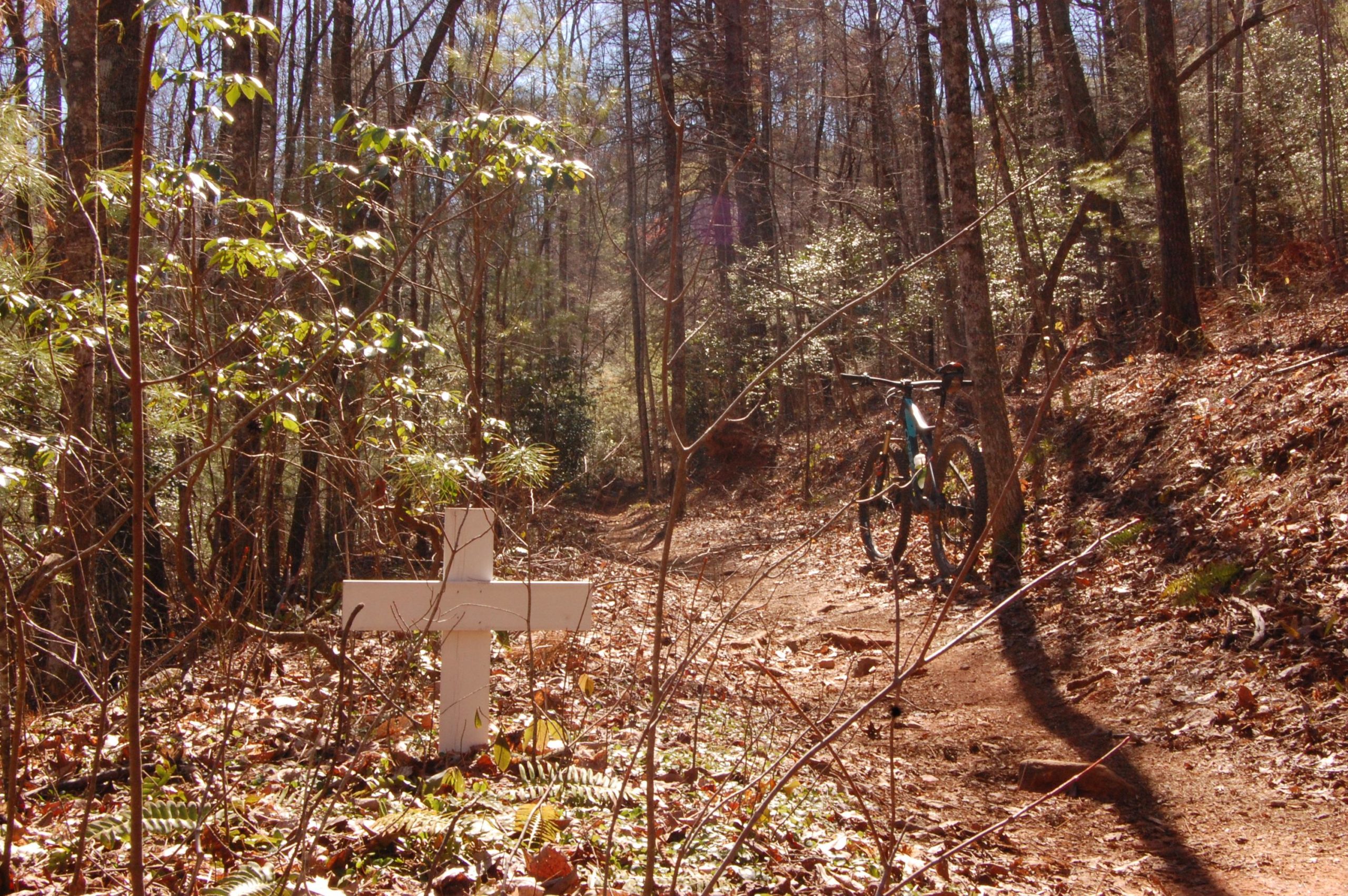 A mountain bike leaning against a tree on a dirt path in a wooded area, with a white wooden cross and fallen leaves in the foreground. Sunlight filters through the trees, illuminating the scene. Jake Mountain Trails mountain bike trail.