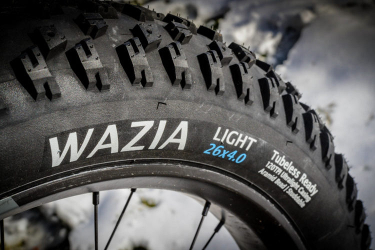 A close-up of a Wazia Light bicycle tire, measuring 26 inches by 4.0 inches, displaying a detailed tread pattern. The tire is labeled as "Tubeless Ready" and features specifications including 120TPI for durability and performance. The background is softly blurred, showing hints of snow.