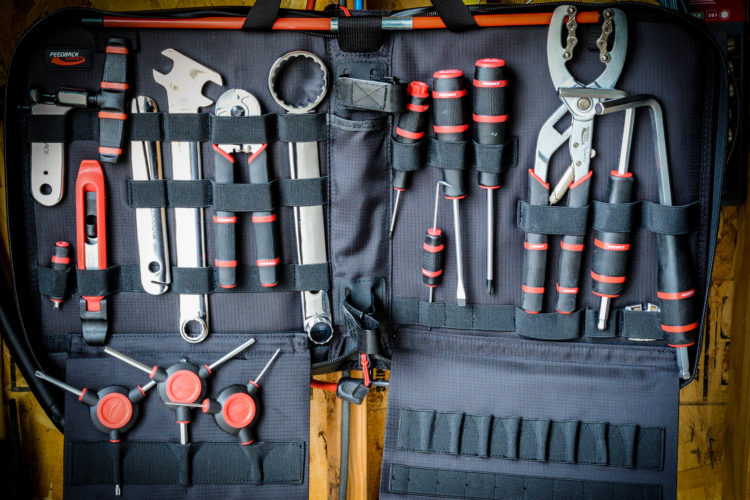 A detailed image of a tool organizer displaying a variety of hand tools, including wrenches, screwdrivers, pliers, and a socket wrench, all arranged neatly in an open fabric case. The tools feature red and black grips and are secured with straps. The background shows a wooden surface with additional wiring and tools.