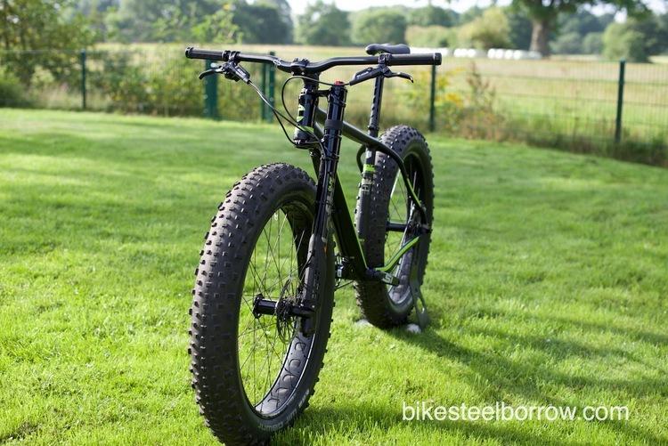 Cannondale Fat CAAD 1: A black fat bike positioned on a grassy area with a backdrop of trees and a fence. The bike features large, wide tires and a sturdy frame, showcasing its design for off-road and rugged terrains.