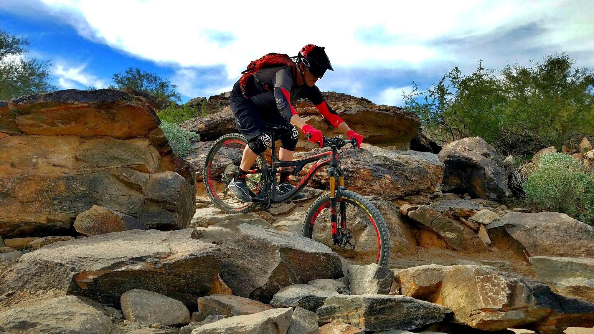 A mountain biker navigating rocky terrain, wearing a helmet, gloves, and protective gear, with a backpack. The scene features large boulders and desert vegetation against a backdrop of blue sky with clouds. South Mountain Park / National Trail mountain bike trail.