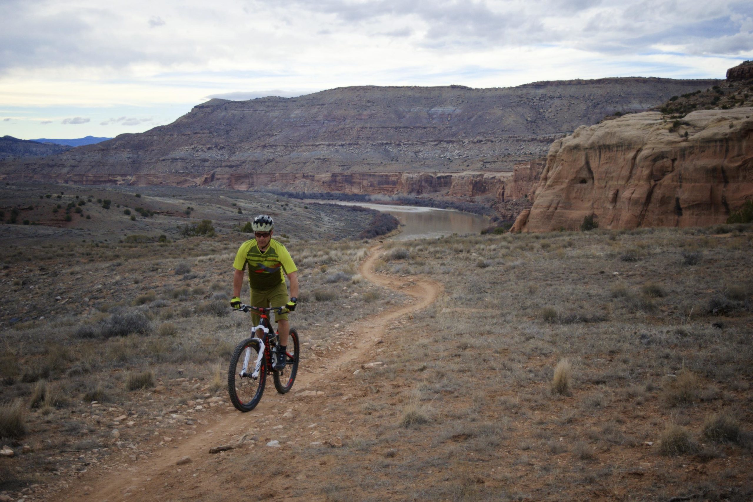 A mountain biker wearing a bright yellow jersey rides along a winding dirt trail in a rugged, arid landscape. In the background, a river flows through a canyon, flanked by steep rock formations and sparse vegetation under a cloudy sky. Kokopelli Area Trails mountain bike trail.