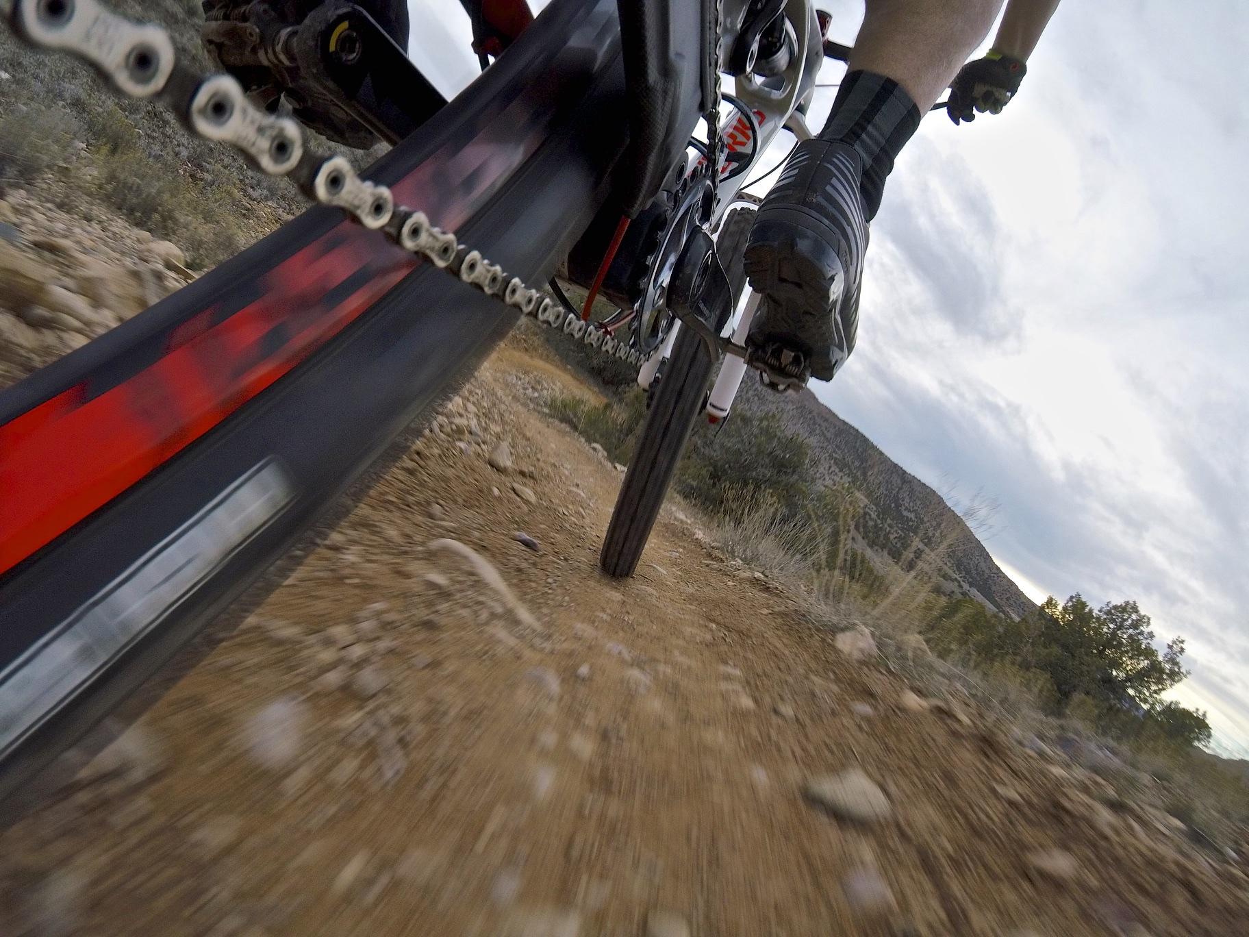 A close-up view of a mountain bike tire and chain while navigating a rocky dirt trail, with a blurred motion effect indicating speed. The background features a hilly landscape and overcast skies. Kokopelli Area Trails mountain bike trail.