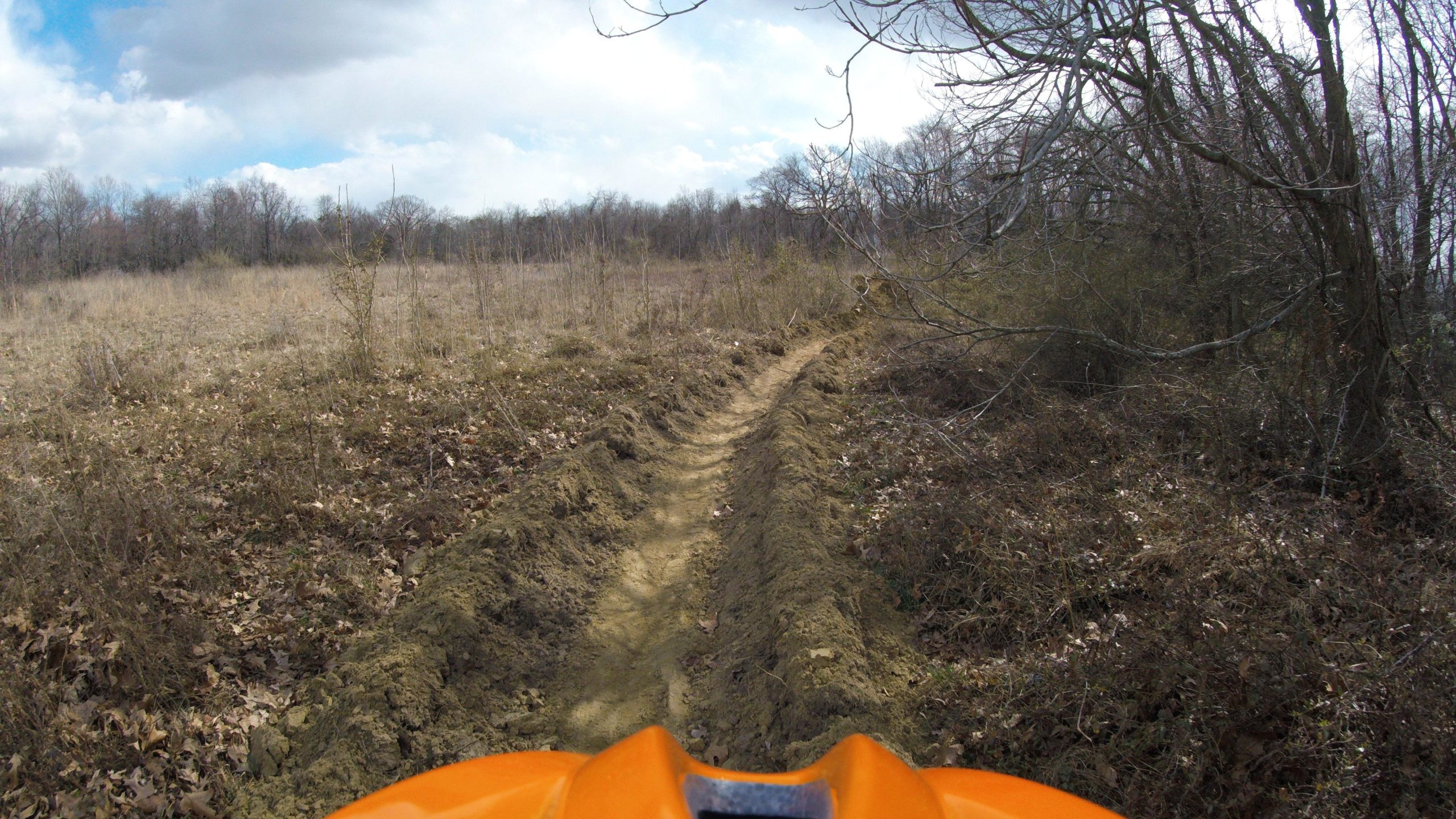 An off-road trail winding through a grassy, open area with sparse trees in the background. The view is taken from the perspective of a rider, showing the upper part of an orange vehicle in the foreground, and a dirt path ahead leading into the wooded area. The sky is partly cloudy. Rancocas State Park - Westampton mountain bike trail.