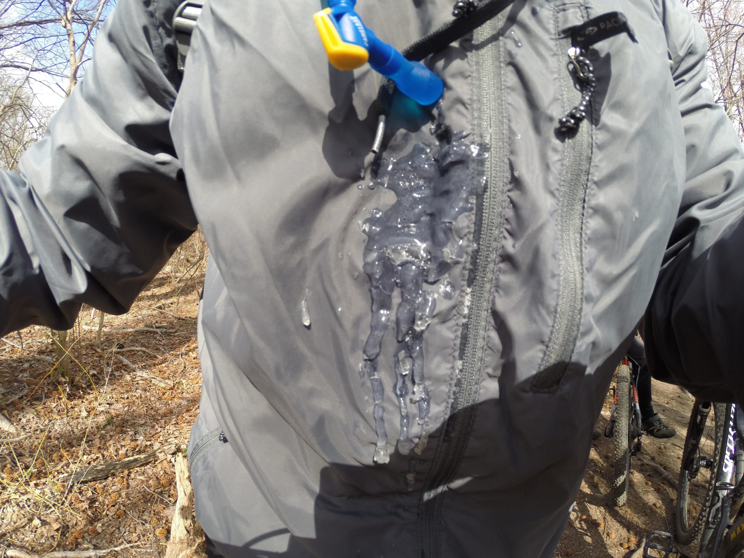 A close-up shot of a person wearing a gray jacket, which features visible water dripping down the front. The jacket is partially unzipped, revealing moisture on the fabric, likely from a hydration pack. The background shows a wooded area with bare trees and some fallen leaves. Bicycles are partially visible on the right side of the image. Richmond Avenue and Forest Hill road mountain bike trail.
