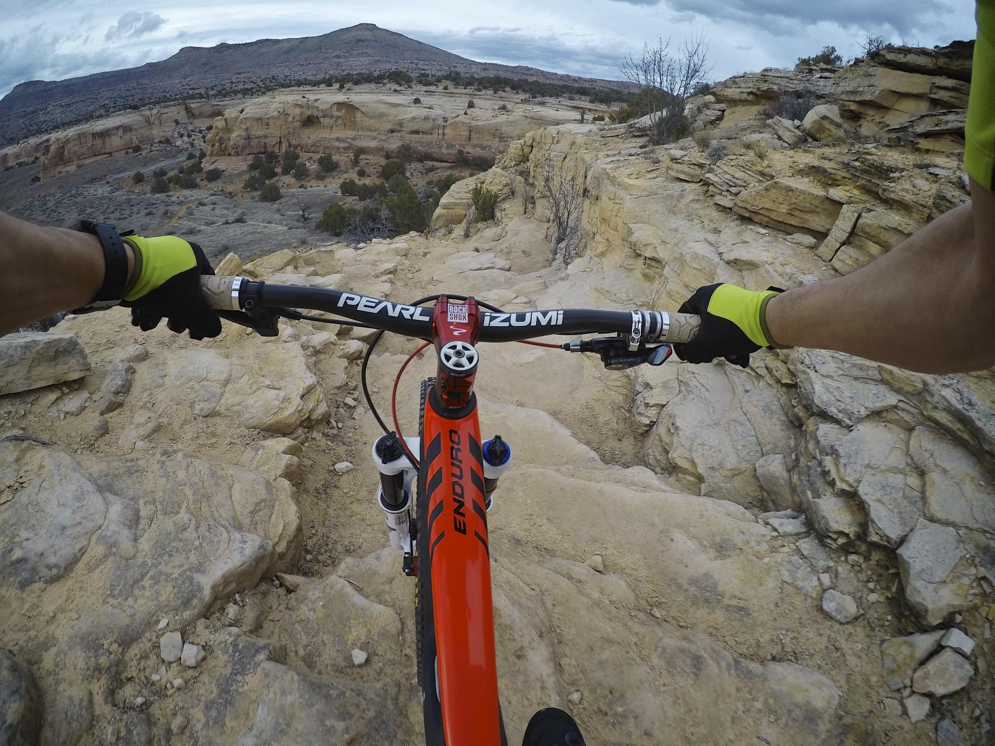 A view from the handlebars of a mountain bike navigating rocky terrain, with a person wearing bright gloves gripping the handlebars. The landscape features layered rock formations and sparse vegetation under a cloudy sky. Kokopelli Area Trails mountain bike trail.