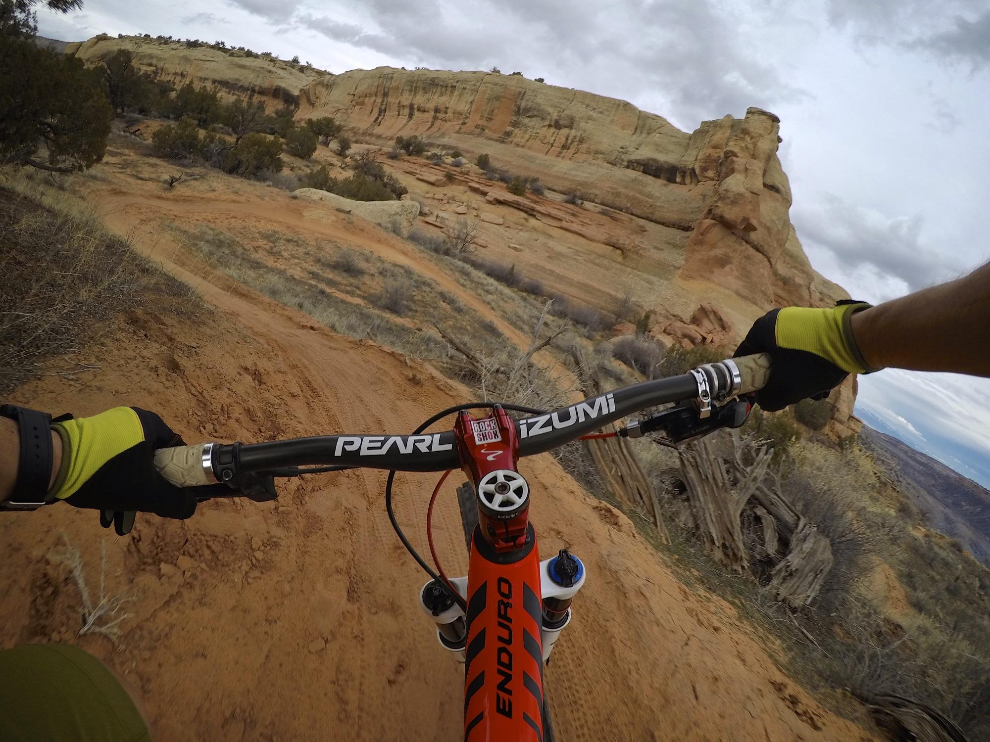 A mountain biker's perspective, gripping the handlebars of an orange bike on a sandy trail, with rocky cliffs and sparse trees visible in the background under a cloudy sky. Kokopelli Area Trails mountain bike trail.