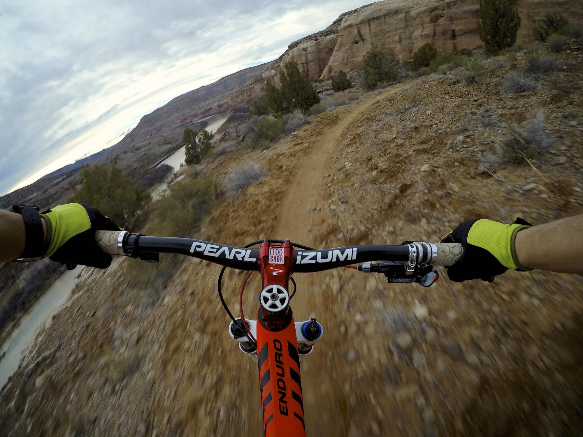 A close-up view of mountain bike handlebars as the rider navigates a rugged trail. The scene features rocky terrain with shrubs and a river visible in the background, under a cloudy sky. The rider is wearing gloves and the bike is equipped with visible components for performance cycling. Kokopelli Area Trails mountain bike trail.