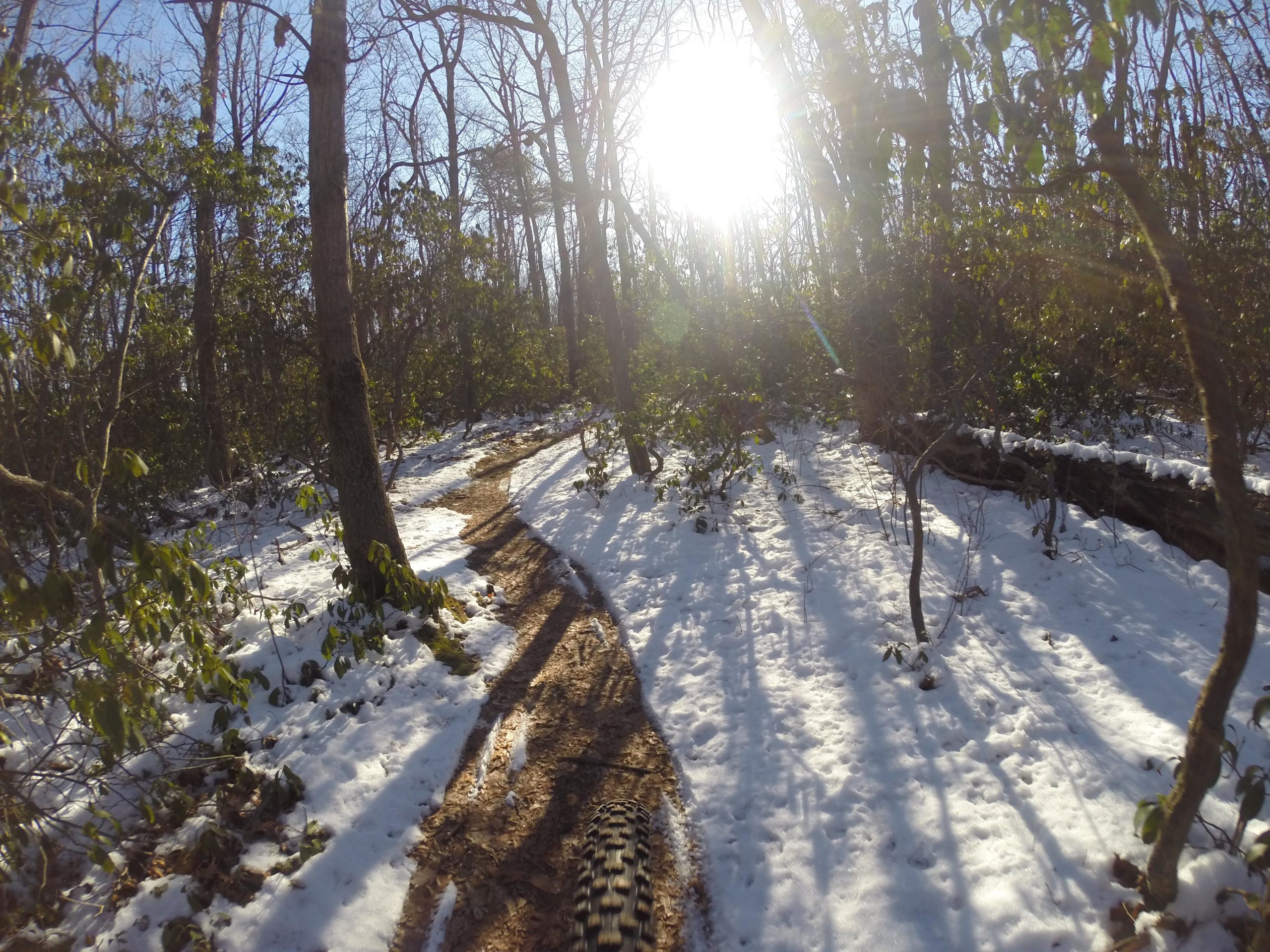 A winding trail through a snowy forest with scattered patches of snow and sunlight filtering through bare trees. The ground shows dirt and tire tracks, indicating the presence of outdoor activity. Allaire State Park mountain bike trail.