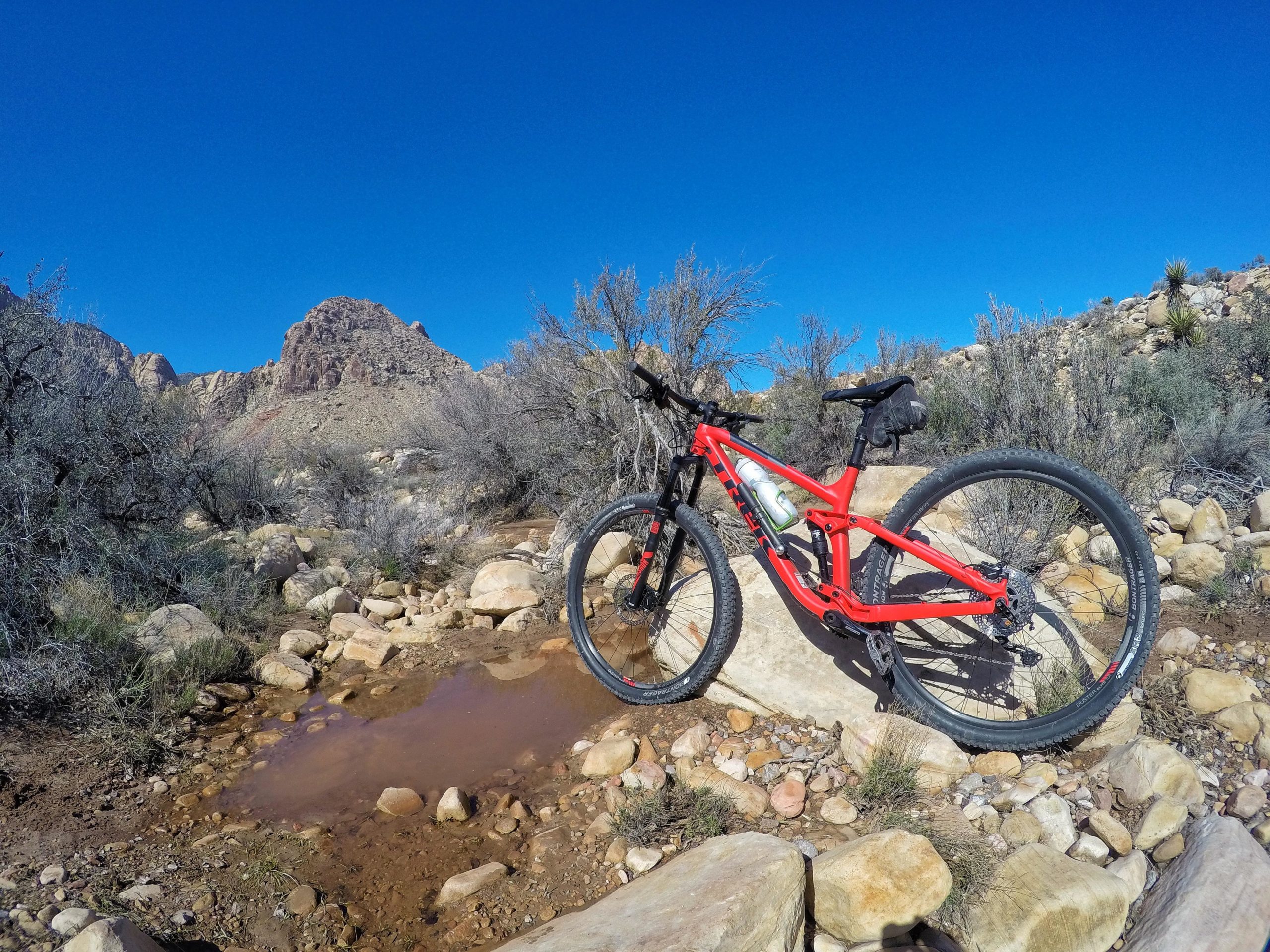 A red mountain bike resting on a large rock beside a muddy puddle, surrounded by desert vegetation and rocky terrain under a clear blue sky. Blue Diamond mountain bike trail.