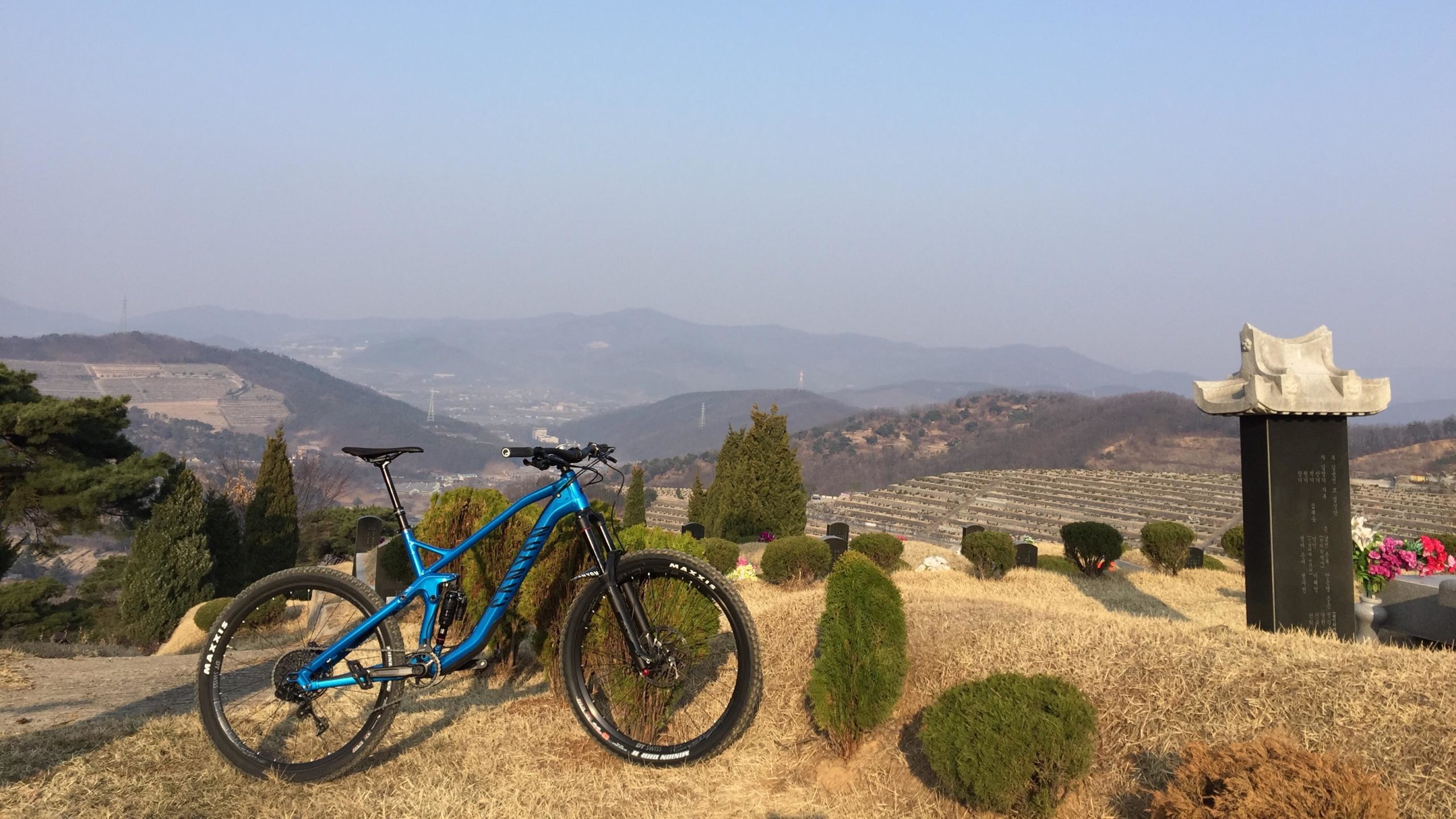 A blue mountain bike is parked on a grassy hilltop surrounded by neatly trimmed bushes. In the background, a landscape of rolling hills and fields is visible under a clear sky. To the right, there is a monument or gravestone with flowers, indicating a serene and reflective atmosphere. Cemetery Ridge mountain bike trail.