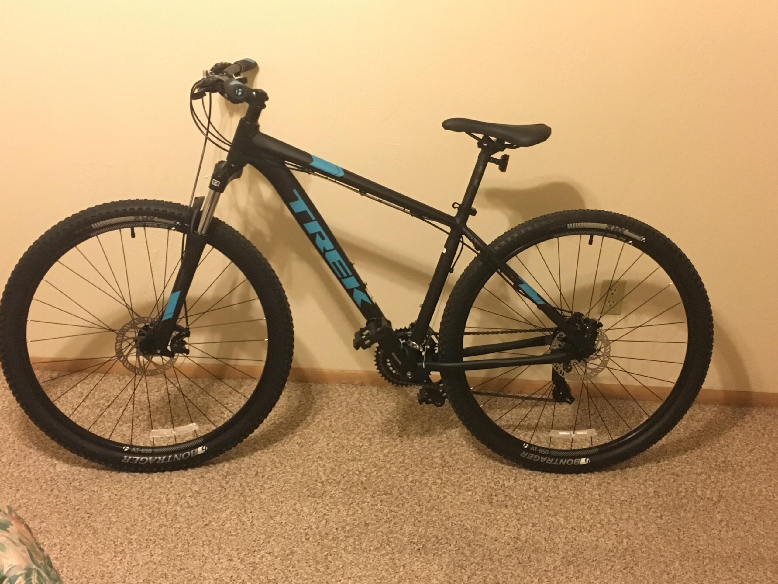 Trek Marlin 5 29er: A black and blue Trek mountain bike leaning against a beige wall. The bike features wide tires suitable for off-road terrain and is positioned on a carpeted floor. The image captures the bike's frame, handlebars, and both front and rear wheels.