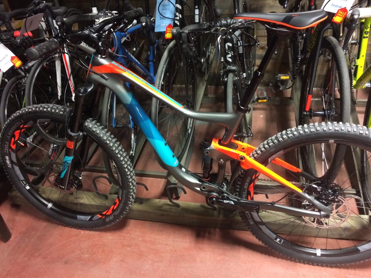 Giant Giant Trance Advanced 27.5 2: A close-up image of a modern mountain bike displayed in a store, featuring a sleek design with a predominantly gray frame accented by blue and orange highlights. The bike is positioned among several other bicycles, showcasing its knobby tires and front suspension fork, indicating suitability for off-road terrain.